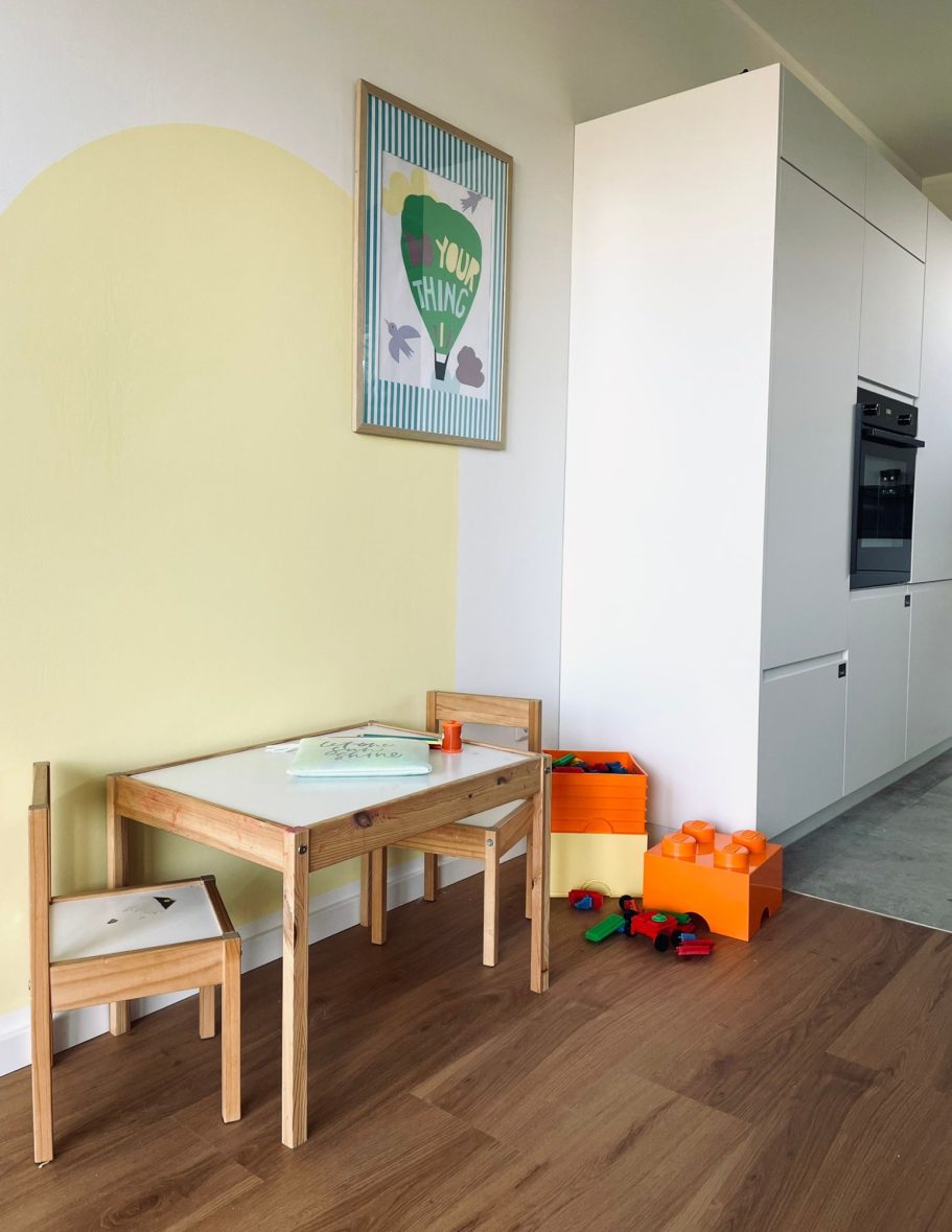 Spielbereich Kindertisch mit Stühlen, bunten Spielzeugkisten und einem Wandgemälde in einem hellen Raum.